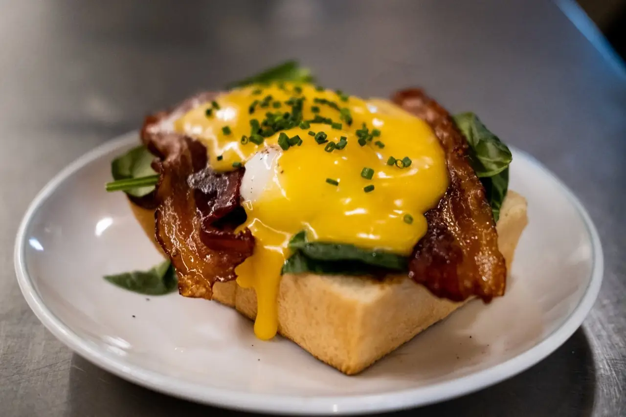 Open-faced brunch toast topped with poached egg, runny hollandaise, crispy bacon, spinach and chives on a white plate