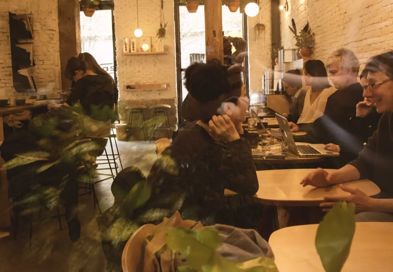 Busy cafe interior seen through the front window, with customers seated at wooden tables working on laptops amid brick walls and hanging plants