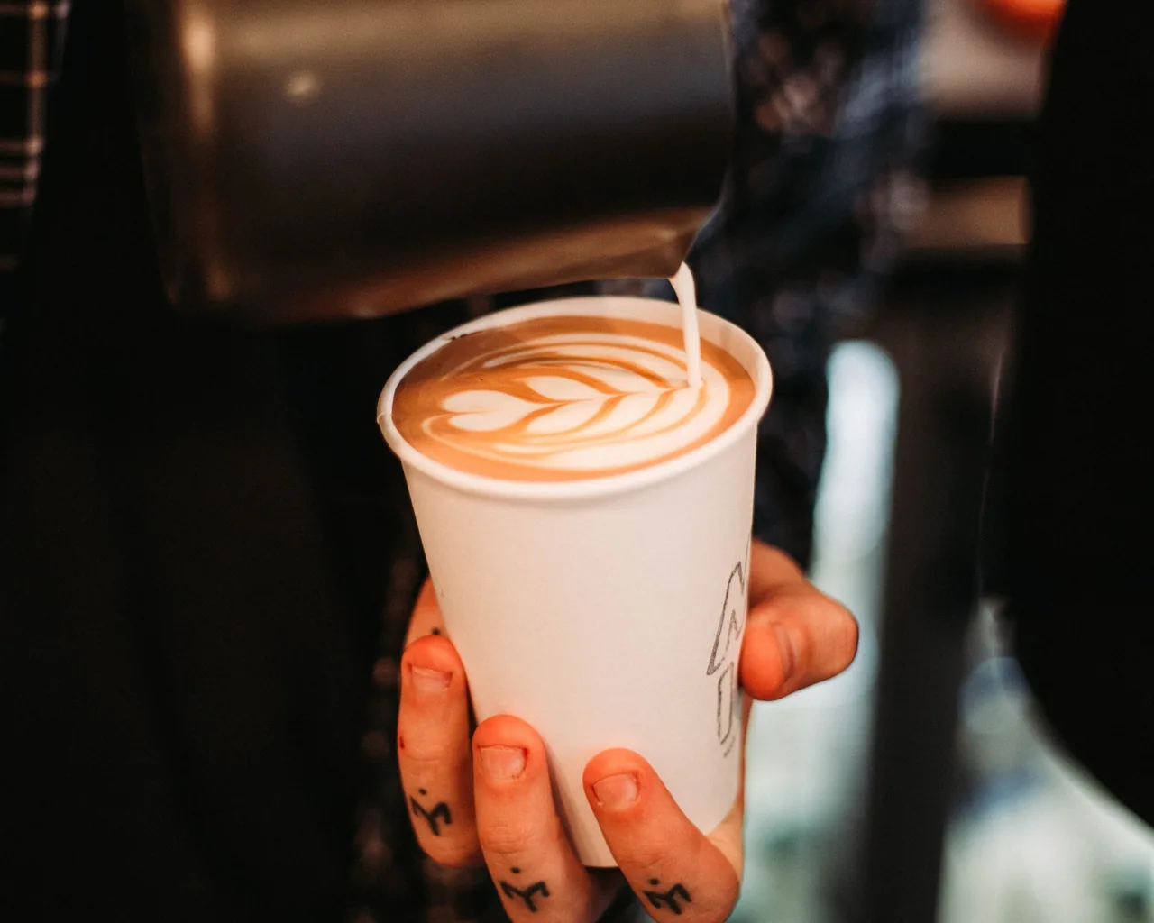 A hand holding a paper cup of latte with a leaf pattern