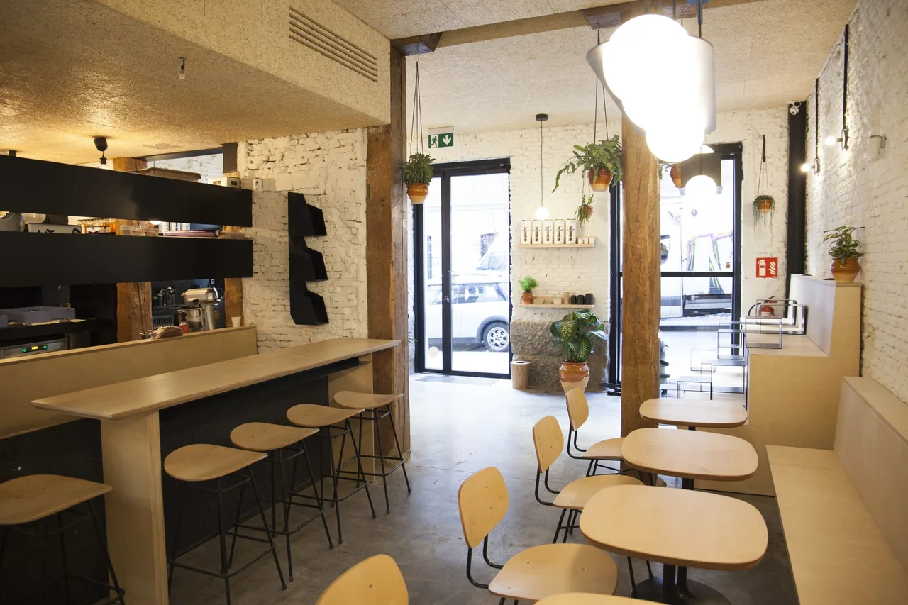 A light cafe interior with a long wooden counter, stools and small round tables