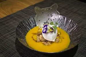 A dessert with a thin crisp tuile over crumble and a scoop of cream on a yellow sauce base, garnished with an edible flower
