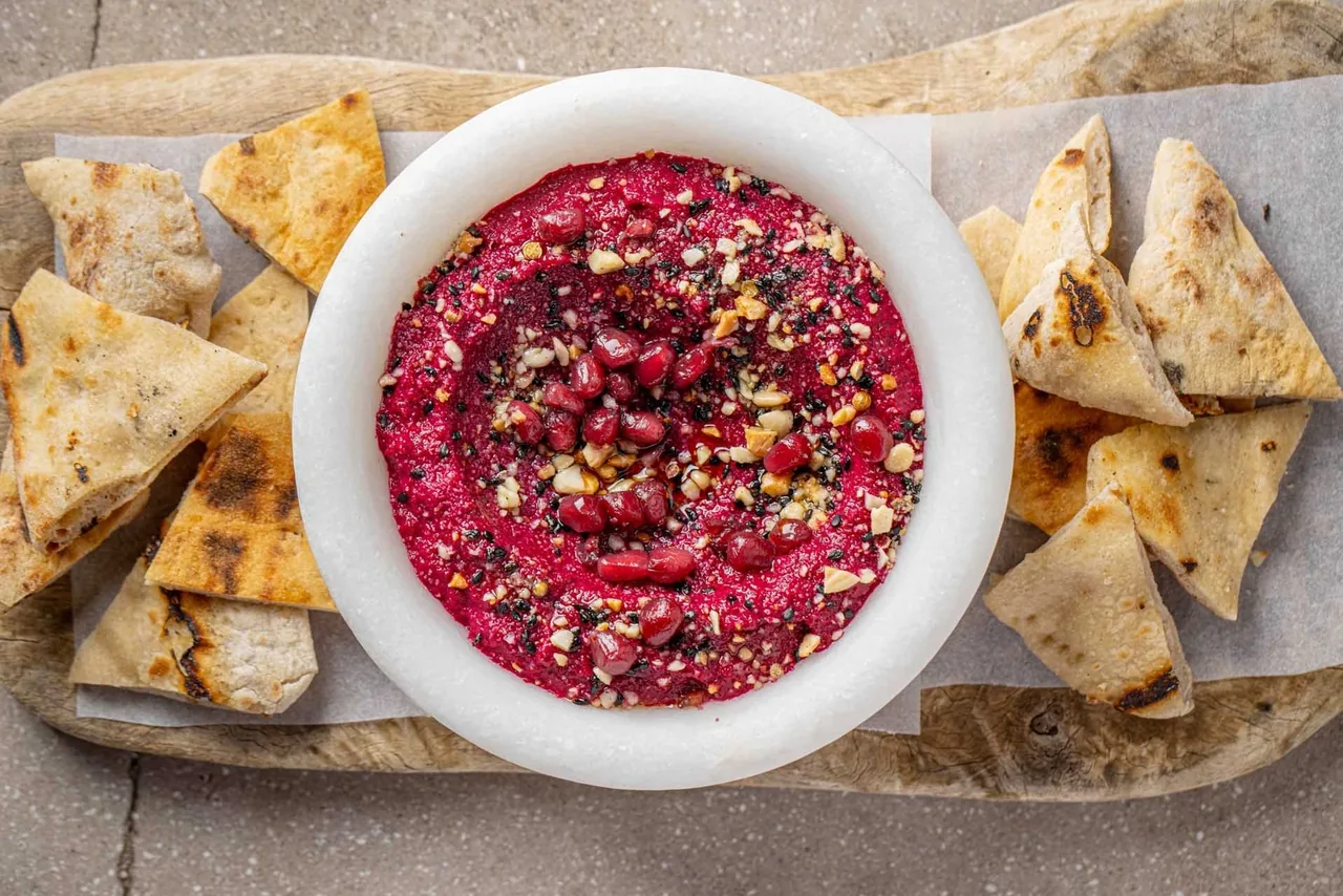 A white bowl of bright pink beetroot dip with flatbread triangles on a board