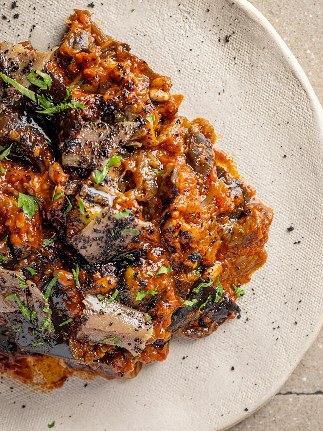 Close-up plated dish of charred eggplant in a rich tomato sauce with pine nuts and chopped parsley