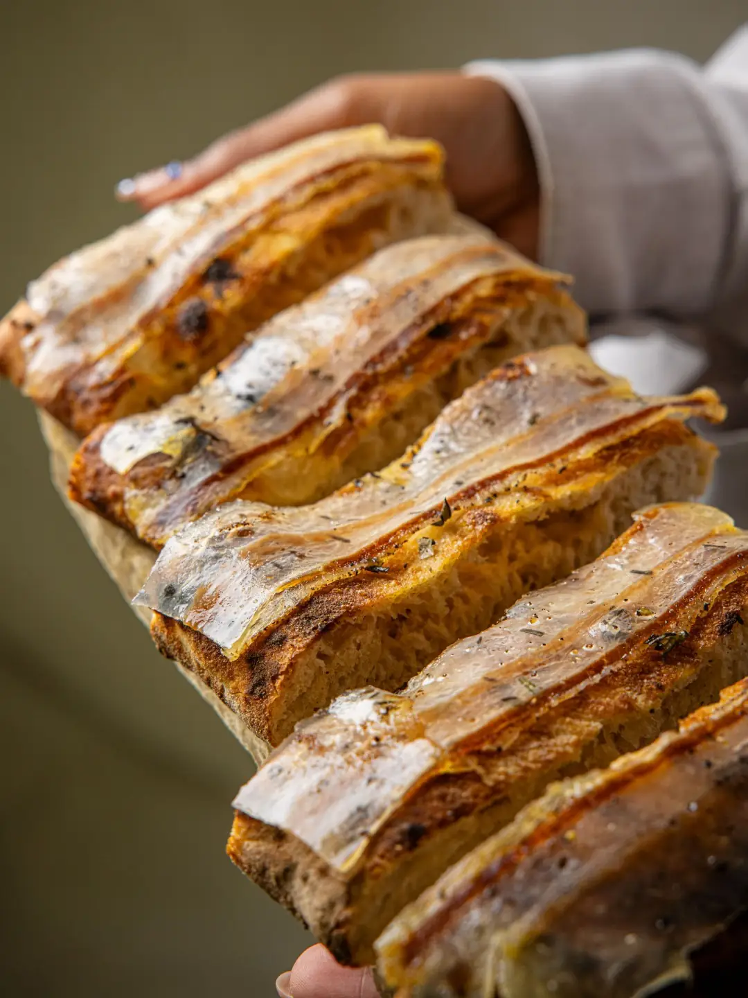 Hands holding a tray of sliced herb-topped focaccia with a crisp, blistered crust
