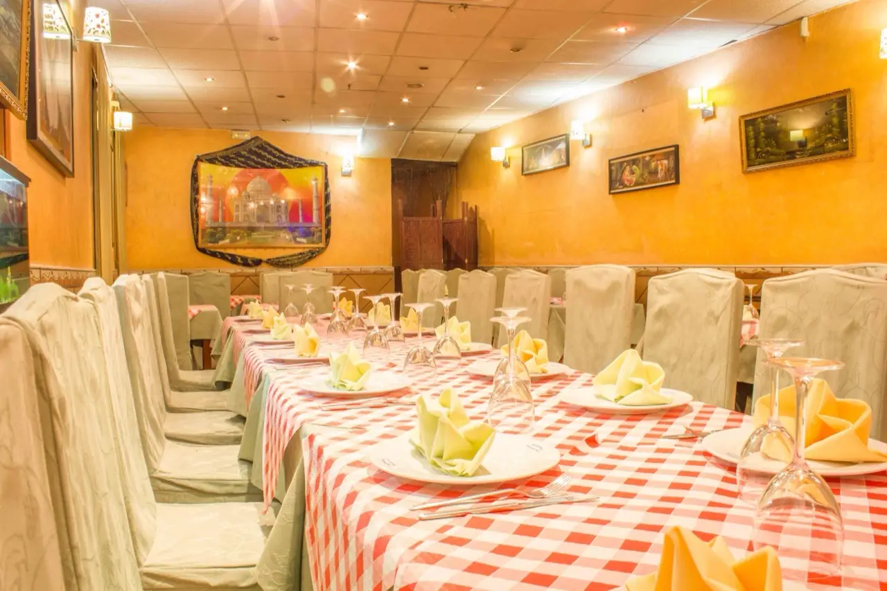 Indian restaurant dining room with ochre walls, framed art, a Taj Mahal picture and long tables set with red gingham cloths and folded napkins