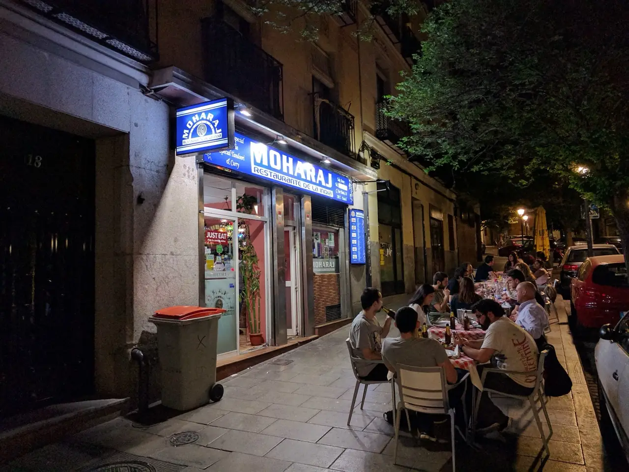 Night exterior of Moharaj Indian restaurant with an illuminated blue sign and diners seated at a row of pavement tables