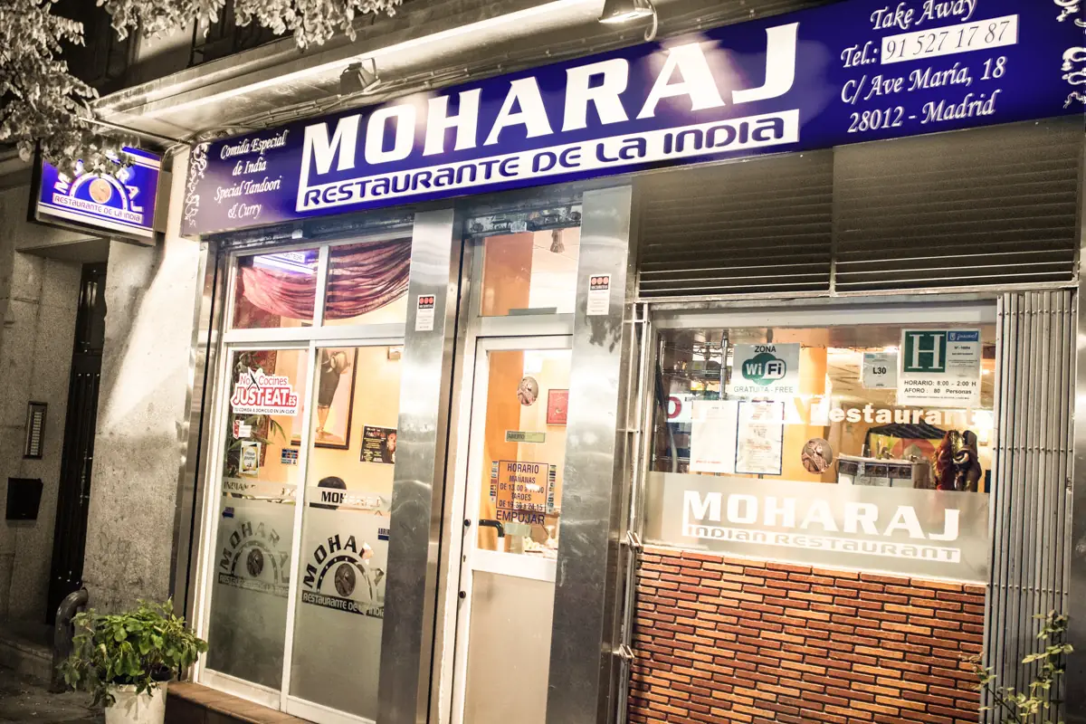 Night close-up of the Moharaj storefront with its large illuminated blue Restaurante de la India sign and glass entrance doors