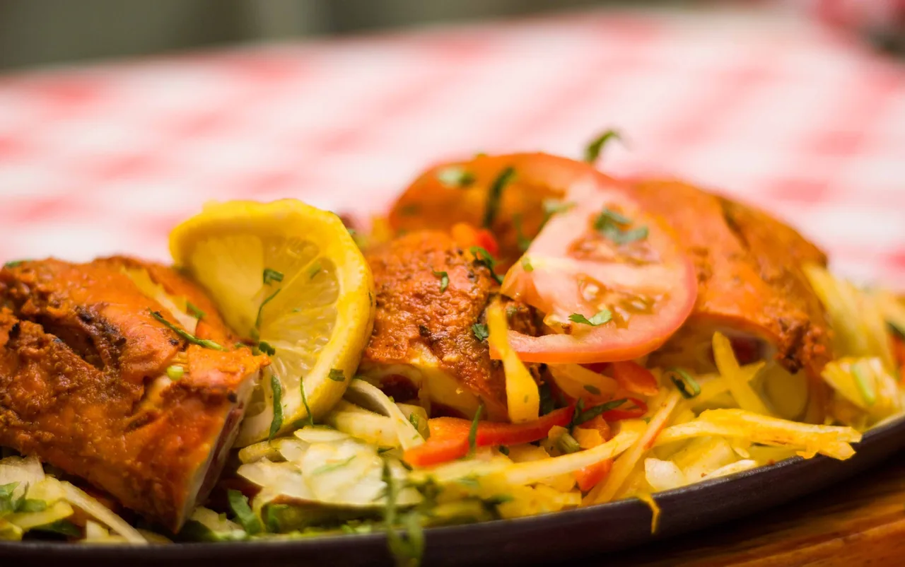 A close-up of red-marinated grilled chicken pieces with a lemon slice, tomato and sliced onions and peppers on a sizzling plate