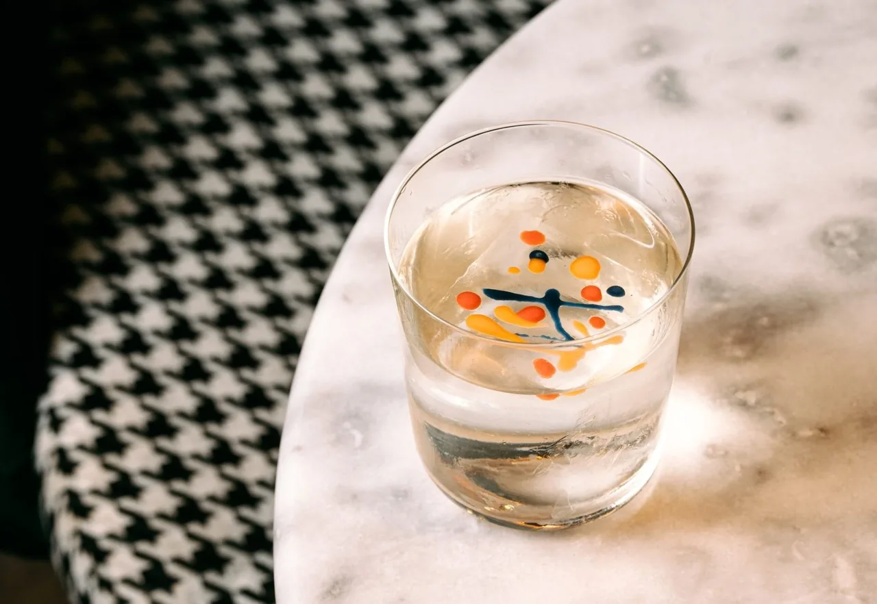 A clear cocktail with an ice cube and small orange and blue markings on a black and white patterned marble table