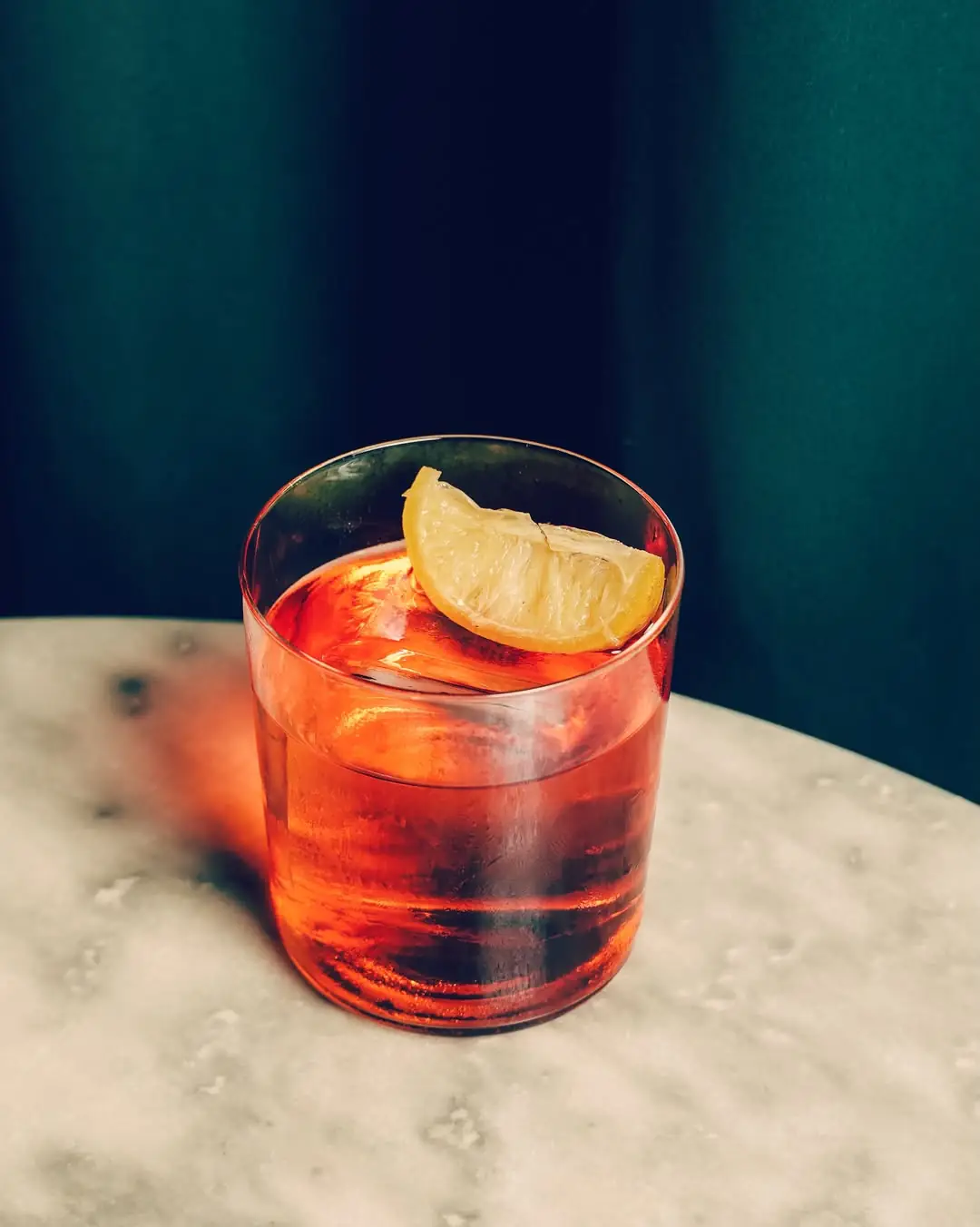 Amber cocktail with a lemon wedge garnish and large ice cube on a marble tabletop