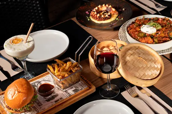 Table spread with a burger, a basket of fries, a glass of red wine, a green cocktail and shared dishes including a saucy chicken plate