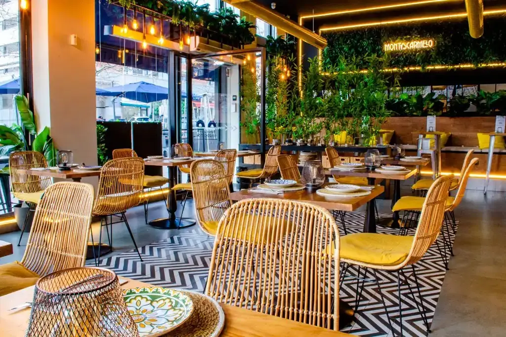 Bright interior with rattan chairs, yellow cushions, hanging bulbs and greenery, a green plant wall with MONTYSCARMELO neon sign and chevron floor tiles