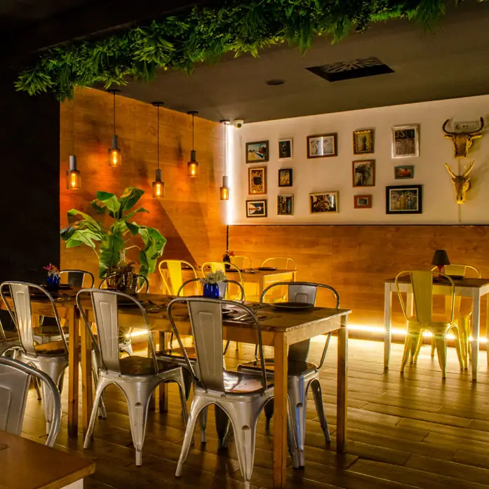 Warm-lit dining room with wooden tables, metal chairs, hanging Edison bulbs, wood-panelled walls with framed pictures and a gilded bull skull