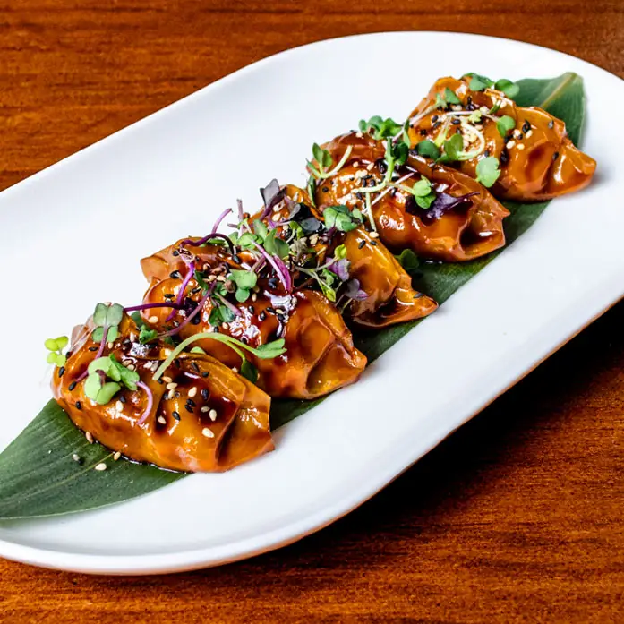 Row of glazed dumplings topped with sesame seeds and microgreens on a green leaf on a white oval plate