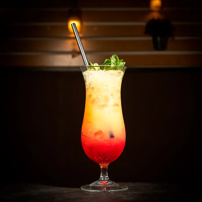 Tall hurricane glass holding a layered red-to-orange cocktail garnished with mint and a metal straw, on a dark bar top
