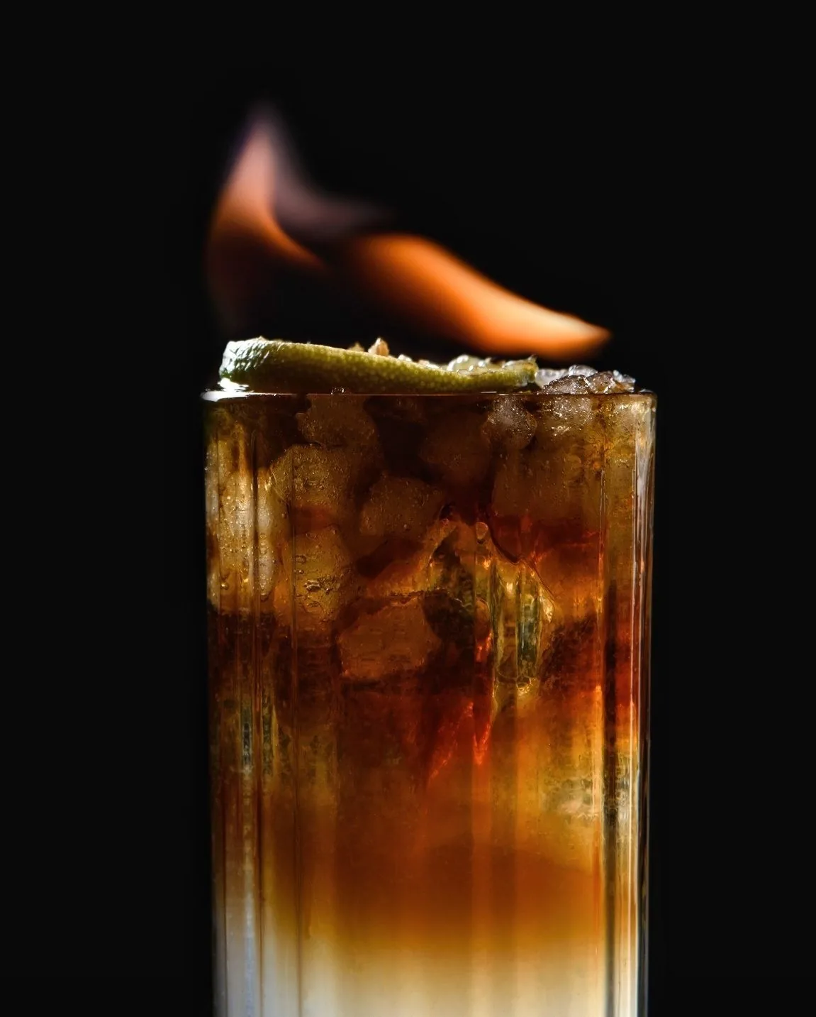 A tall drink with ice and a lit flame on top against a black background