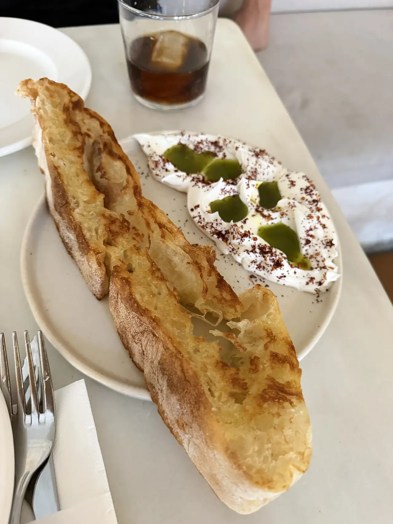 Toasted bread with whipped cream cheese dip topped with green oil and sumac, and a cold drink on the table