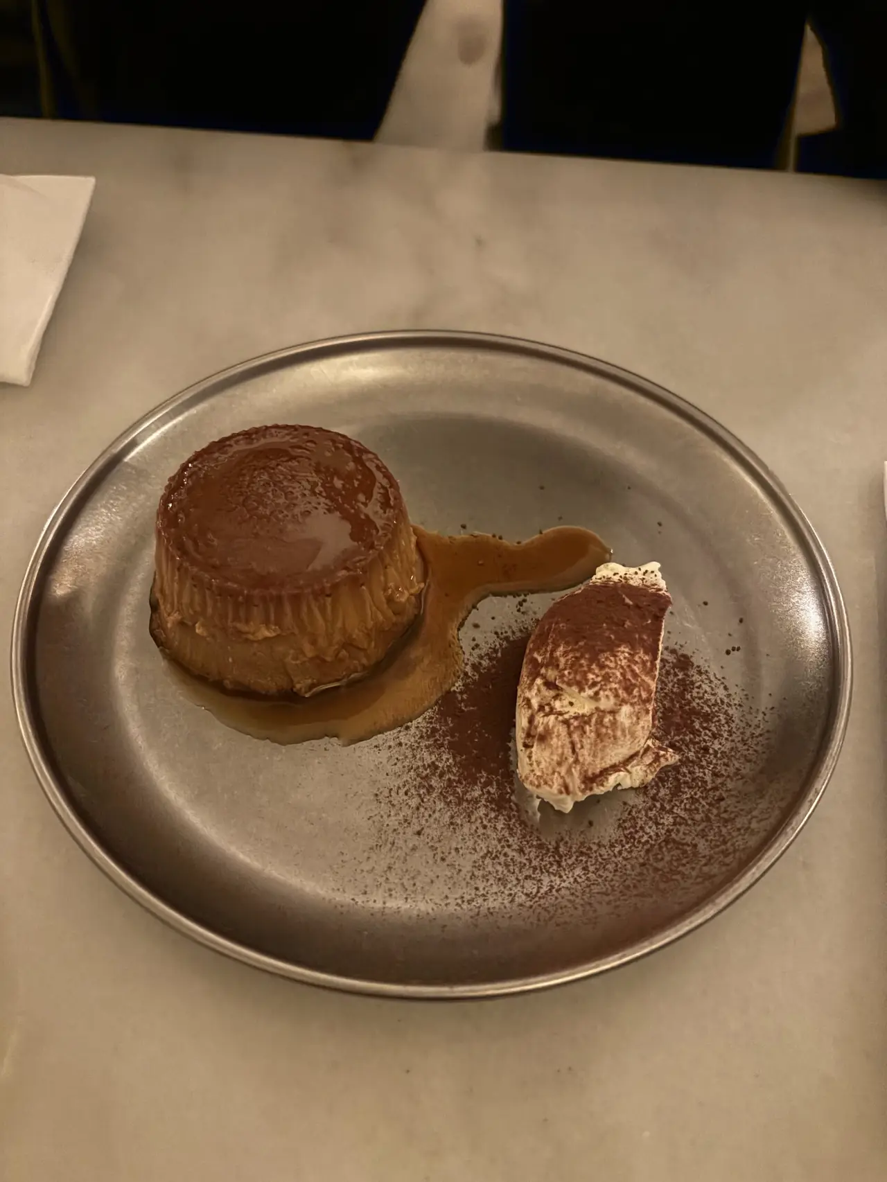 Flan with caramel sauce next to a cocoa-dusted whipped cream quenelle on a metal plate