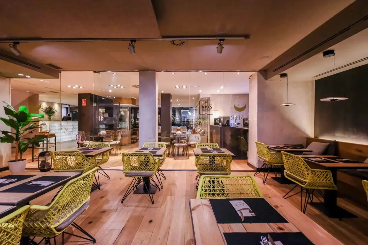 Nadir restaurant dining room with wooden floor, set tables and bright yellow lattice chairs, the Nadir/Zenit Hoteles glass signage toward the lobby (image mirrored)