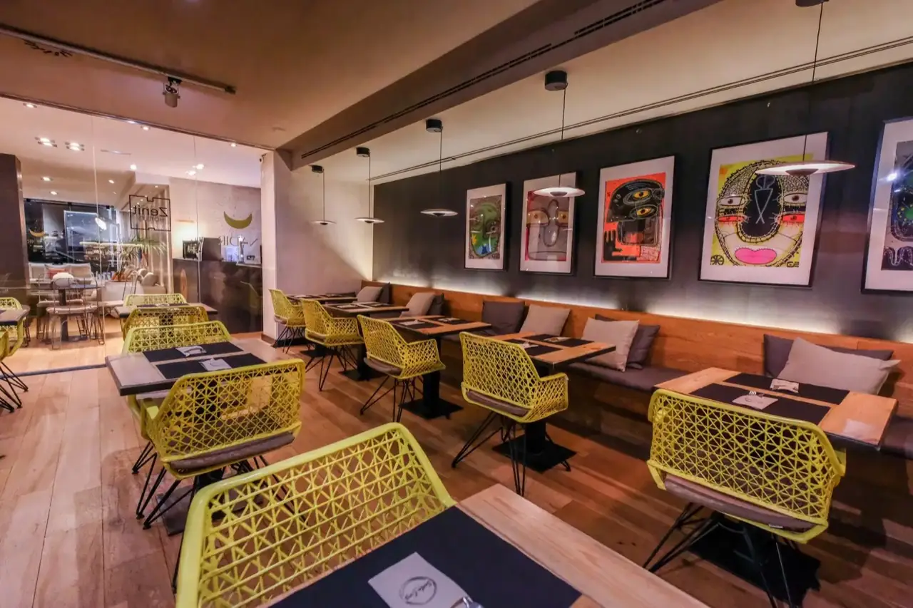 Dining room with yellow lattice chairs, a wooden banquette with cushions and a row of colourful framed face-motif artworks on a dark wall