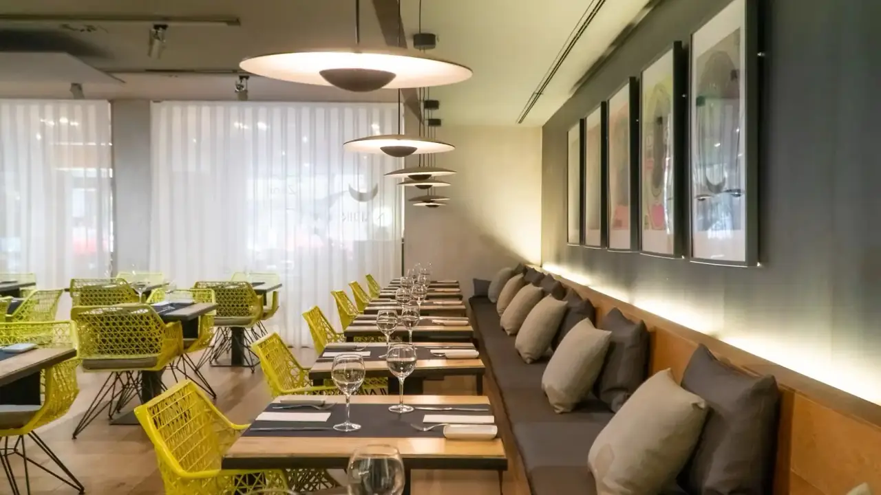 Set banquette tables with wine glasses and dark placemats under saucer pendant lights, framed prints along the wall