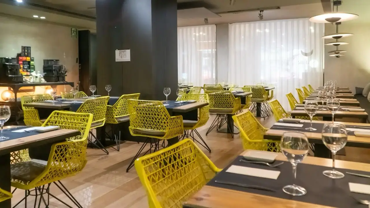 Dining room of set tables with wine glasses and yellow lattice chairs, a coffee-machine station and sheer curtains at the window