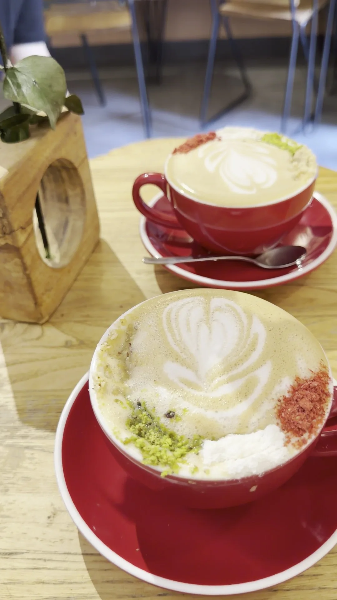 Two cups of latte with art in red cups and saucers, one topped with pistachio and pink crumbs