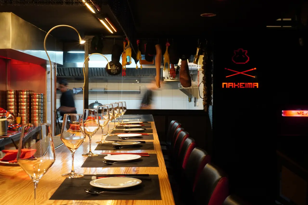 A long pine counter laid with dark mats, white plates, red chopsticks and rows of wine glasses, facing an open kitchen where cooks work; rubber chickens, a cured ham leg and other props hang from a rail overhead, and a red neon sign reading NAKEIMA above crossed chopsticks glows on the black wall at the right.