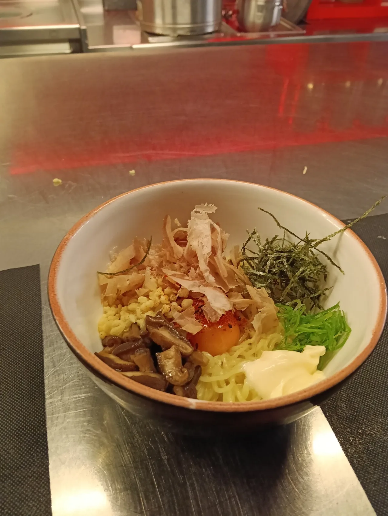 A small bowl of noodles topped with bonito flakes, shredded nori, a raw egg yolk and a spoonful of mayonnaise
