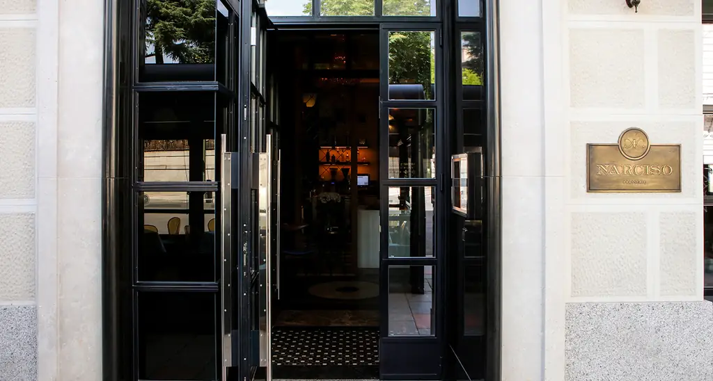 Entrance of Narciso with open black-framed glass doors and a brass 'NARCISO' plaque on the pale stone facade