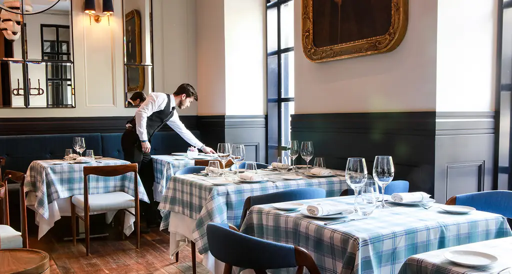 Dining room with blue and white plaid tablecloths, blue banquettes and wood chairs, a waiter setting a table by tall windows