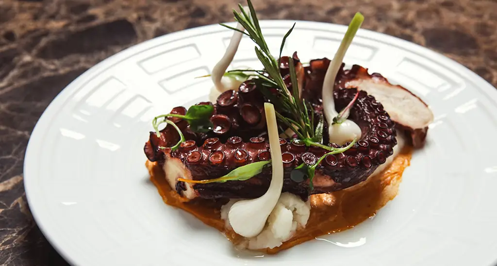 Grilled octopus tentacle over potato puree with baby spring onions, herbs and a smear of red sauce on a white plate