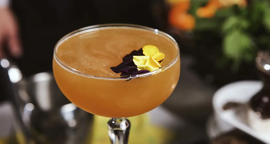 Amber cocktail in a coupe glass garnished with yellow and purple edible flower petals