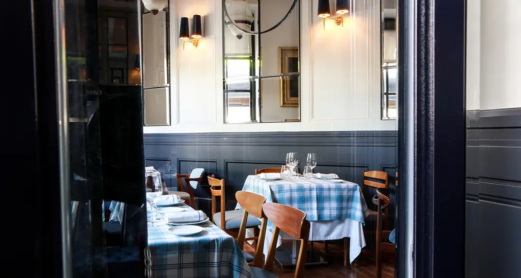 Dining room corner with plaid tablecloths, wood chairs, dark wainscoting, framed mirrors and wall sconces
