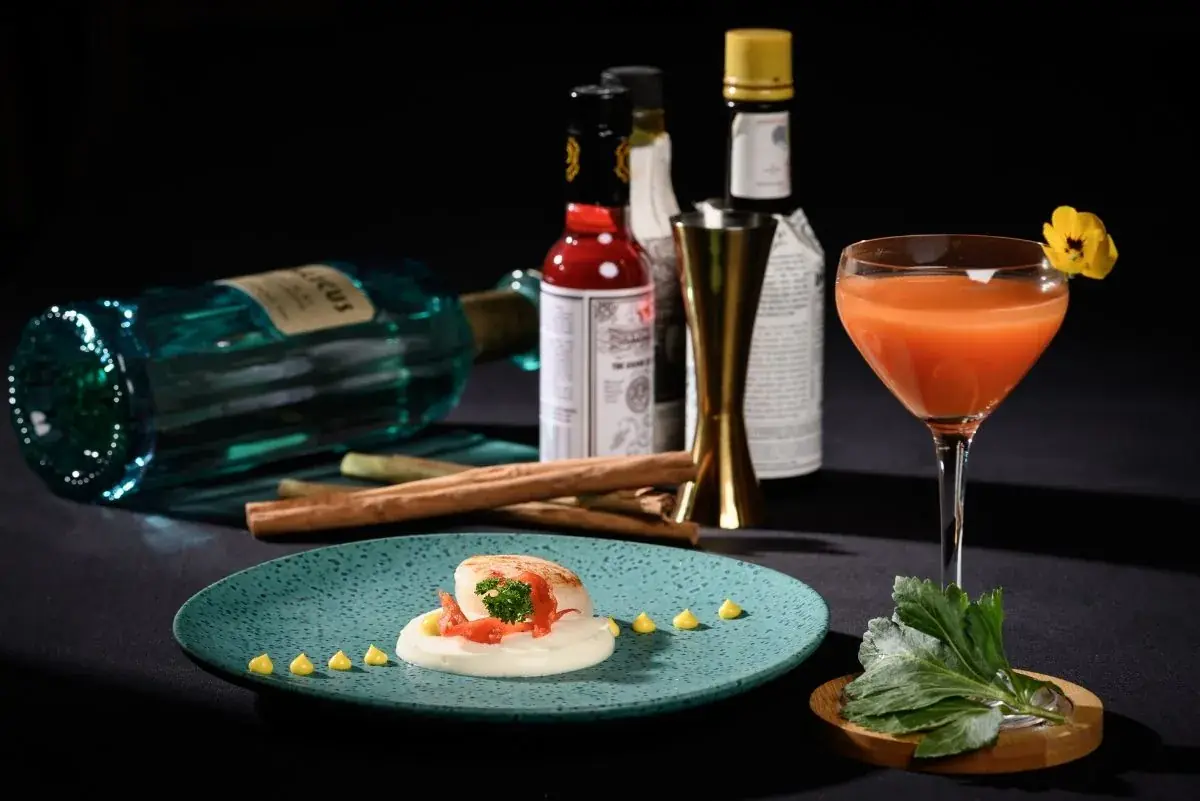 Seared scallop on a swirl of sauce with a citrus dressing, plated on a teal ceramic dish beside an orange cocktail, at Negresco Princess Rooftop