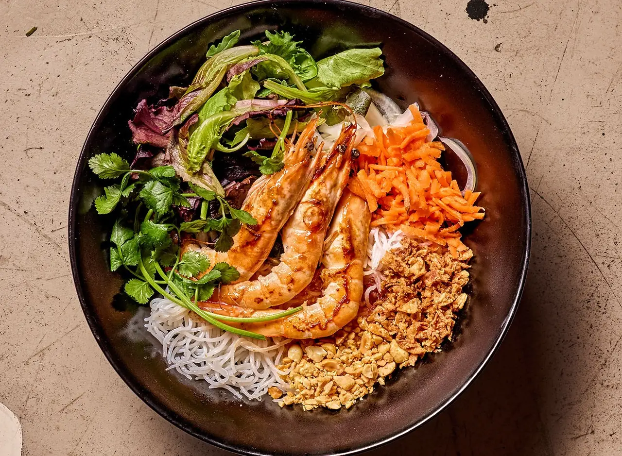 Vietnamese vermicelli bun bowl with grilled prawns, shredded carrot, crushed peanuts, fried shallots and fresh herbs in a black bowl