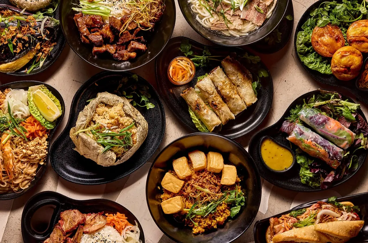 Overhead spread of Vietnamese dishes including pho, grilled spring rolls, fresh summer rolls, fried tofu rice and salads on black plates