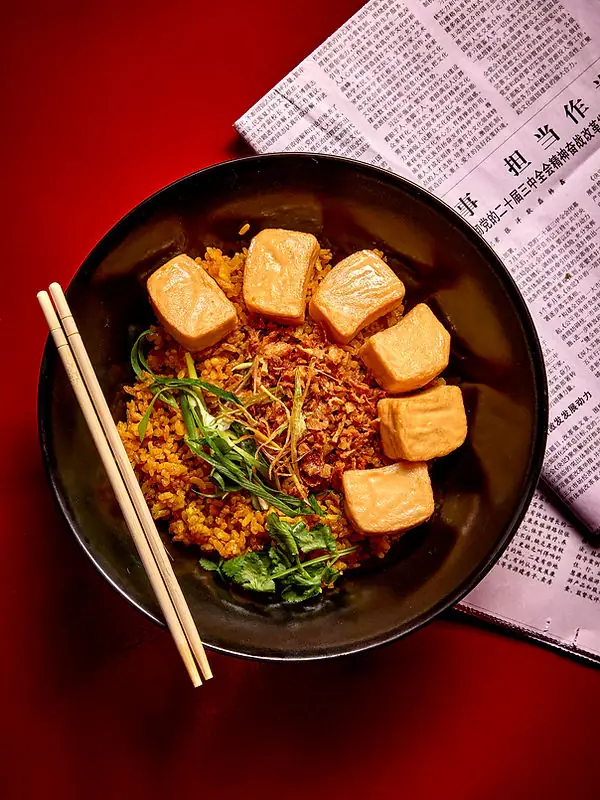 Black bowl of fried rice topped with golden tofu puffs and herbs, chopsticks resting alongside on a red surface