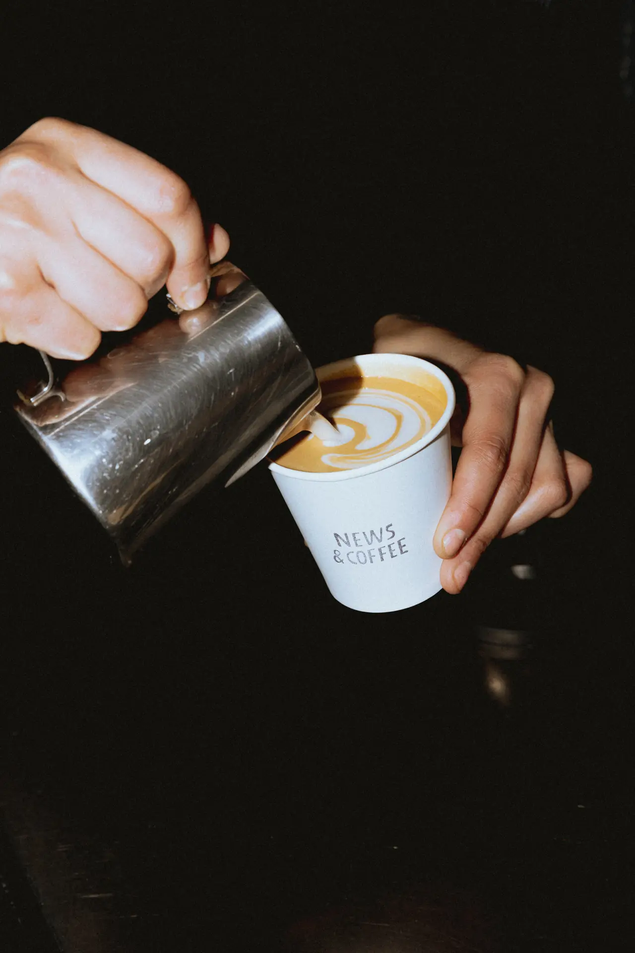 Barista pouring steamed milk into a paper cup marked News & Coffee, creating latte art in a golden-brown espresso drink