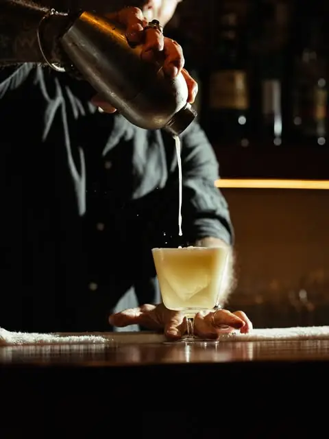 Bartender straining a pale frothy cocktail from a metal shaker into a coupe glass on a bar mat in dim light