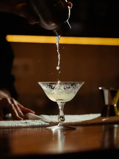 Cocktail being strained from a shaker into a stemmed coupe glass on a bar mat with a warm backlight