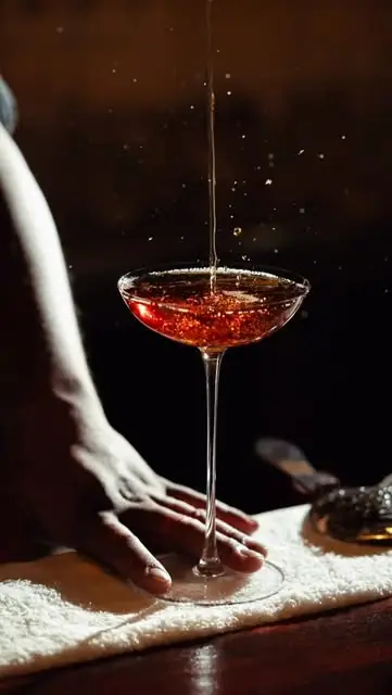 Deep red cocktail in a tall coupe glass being finished with a pour on a wooden bar top