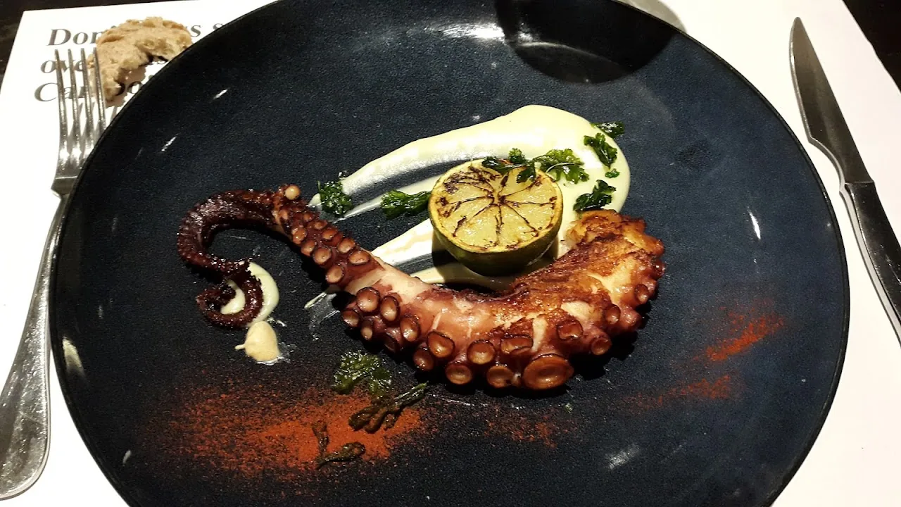 Grilled octopus tentacle on a black plate with citrus and sauce dots