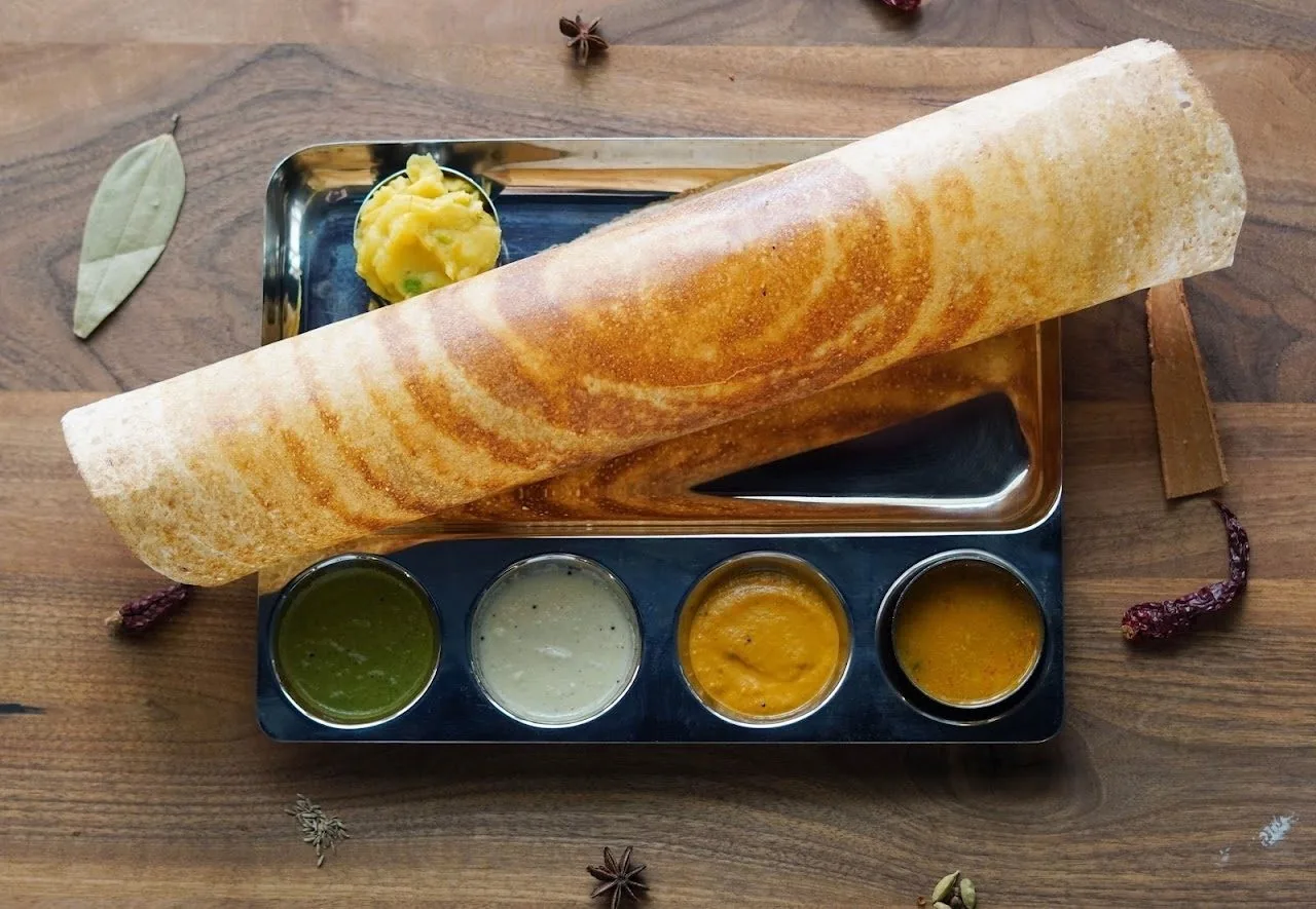 A long rolled dosa on a steel tray with a small portion of potato and four small bowls of chutney and sambar