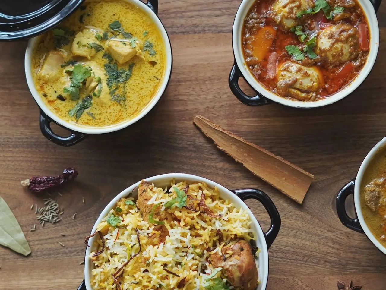 Overhead view of black and white handled pots holding a yellow curry, a red curry and a rice dish, with whole spices on a wooden table