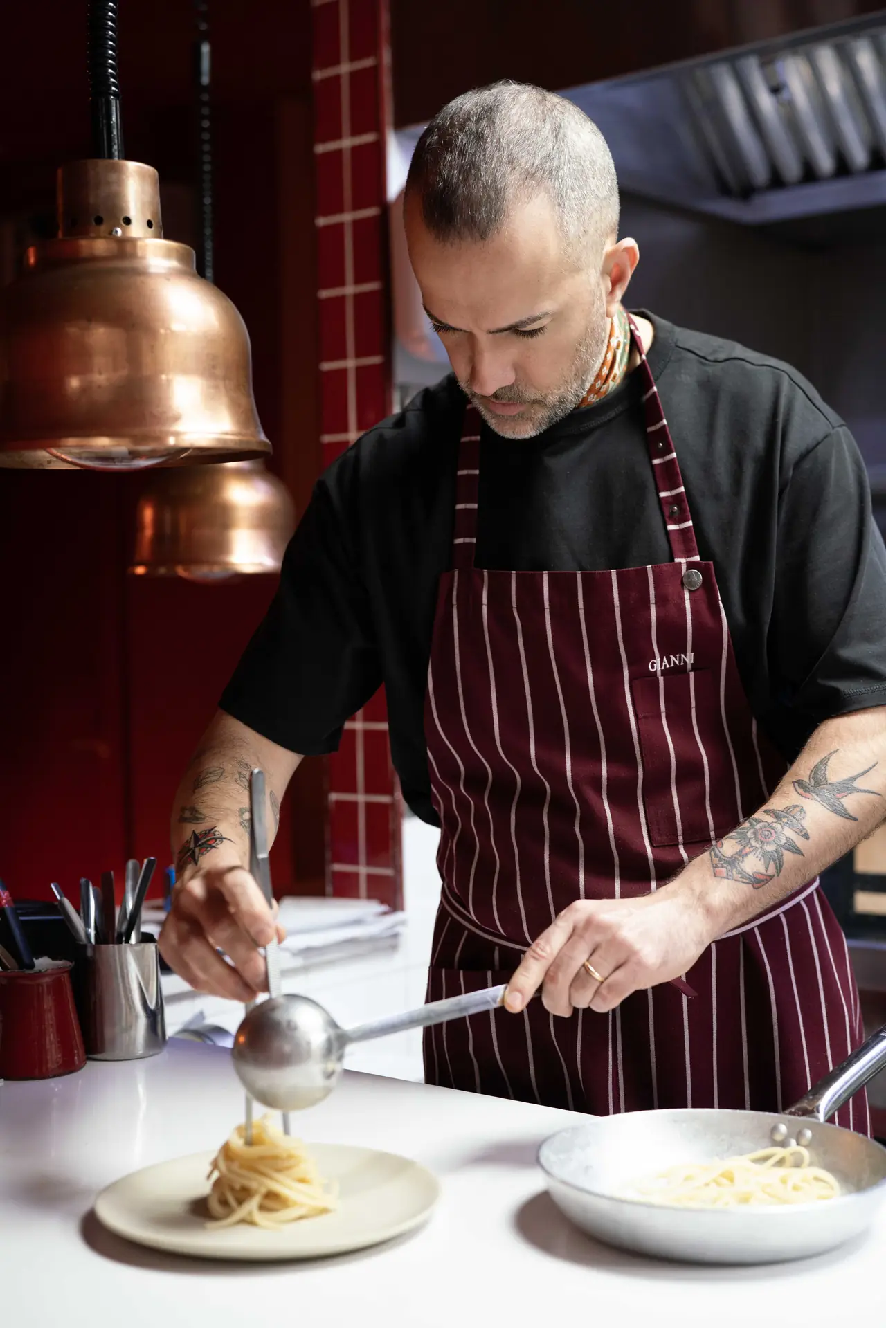 Chef in a burgundy pinstriped apron holding a ladle over a nest of spaghetti plated on a pale dish, a pan of pasta beside it on the white kitchen pass under a copper heat lamp
