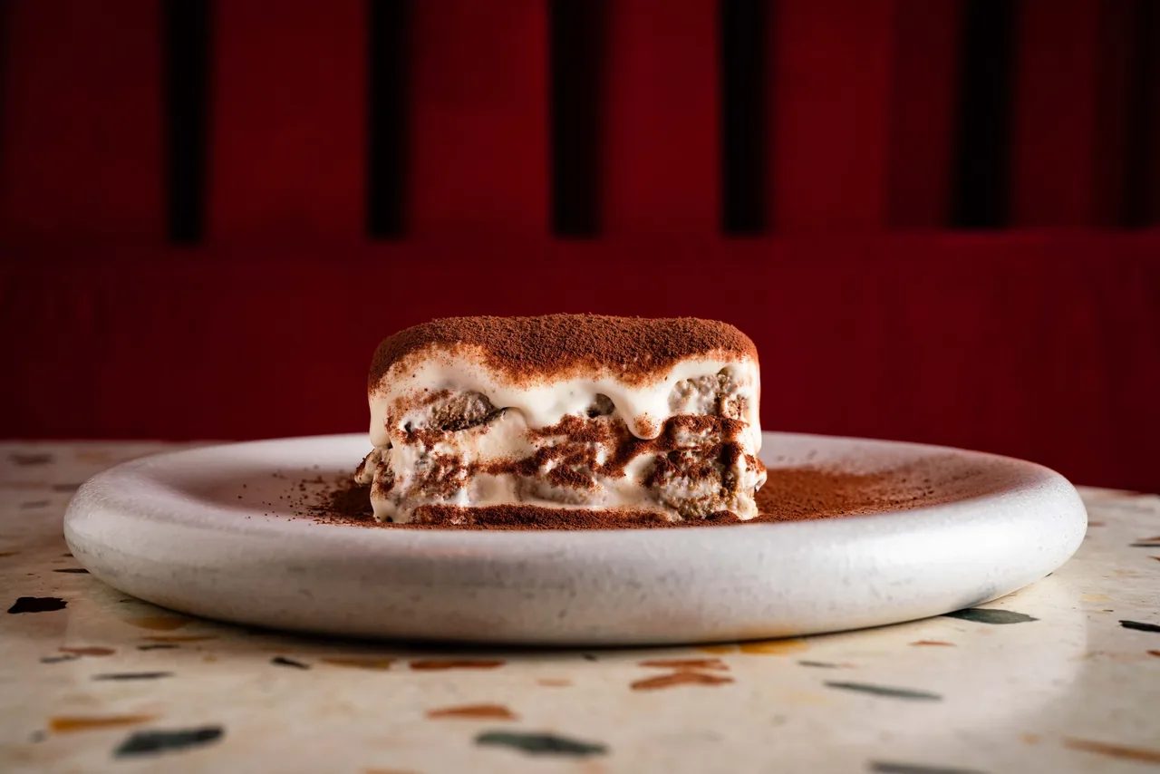 A slice of tiramisu dusted with cocoa on a round white plate in front of a red velvet seat