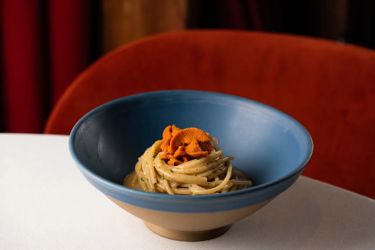 A blue bowl of spaghetti topped with orange sea urchin, in front of a red velvet chair