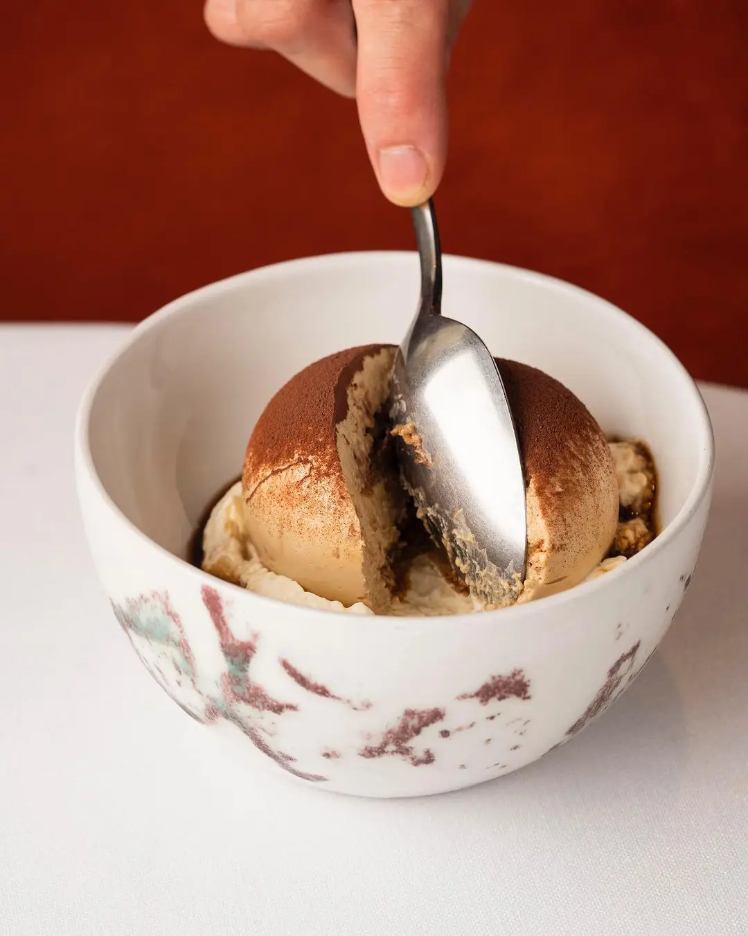 Spoon cutting into a tiramisu-style dessert dusted with cocoa in a marbled bowl