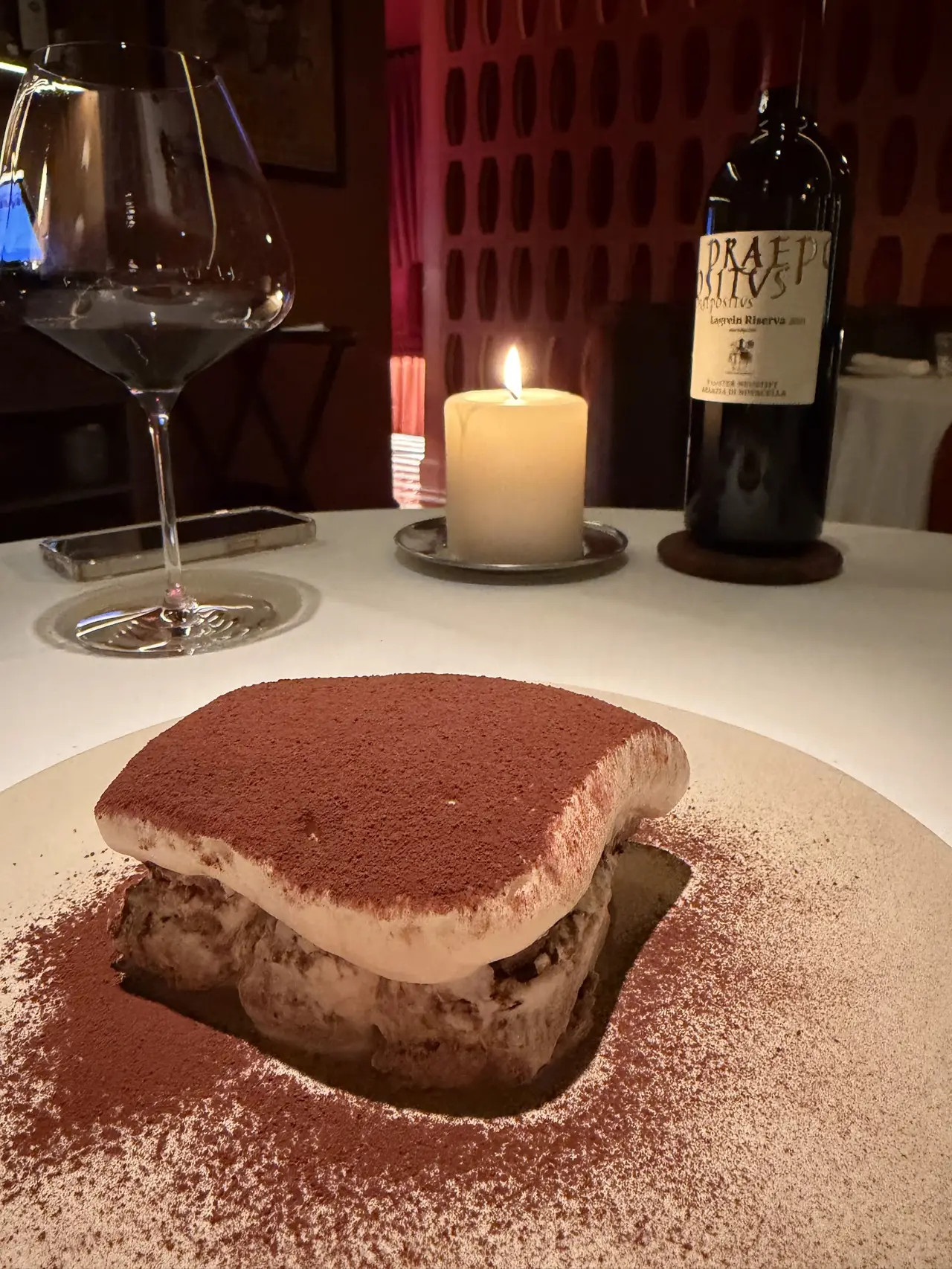 Slice of tiramisu dusted with cocoa on a plate, wine bottle and candle in background